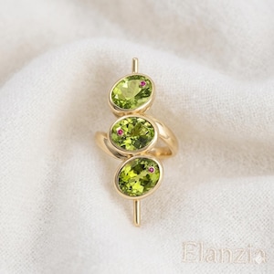 May include: A gold ring featuring three oval-cut peridot gemstones, each accented with a small, round ruby. The peridot stones are a vibrant green, set in a swirling design. The ring is displayed on a white, textured surface.