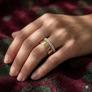 May include: Two gold rings on a finger. One ring is a wide band, and the other is a thinner band with small, clear stones. The rings are set against a burgundy and green patterned background.