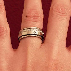 May include: A silver ring with a band of small, clear gemstones encircling the top. The ring has a wider band with a polished finish and a series of small, circular indentations. The ring is on a hand with a red background.