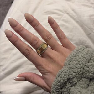May include: A gold-colored ring with a wide band and a row of small, clear stones. The ring is on a finger with a French manicure. The ring has a rectangular shape with a band of small stones.