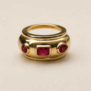 May include: A wide gold band ring featuring a central rectangular red gemstone flanked by two smaller teardrop-shaped red gemstones. The polished yellow gold metal has a smooth, rounded surface with the three stones set securely into the band.