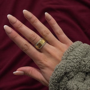 May include: A gold-colored ring with a wide band and a row of small, clear stones. The ring is on a hand with French-tip nails. The background is a deep red color.