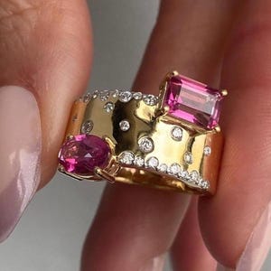 Puede incluir: Un anillo de banda ancha de oro con una piedra preciosa rosa de corte esmeralda rectangular y una piedra preciosa rosa de corte ovalado. La superficie de la banda de oro está decorada con numerosos diamantes blancos pequeños, redondos y claros.