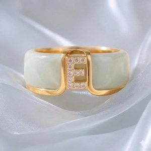 May include: A gold ring with a wide, pale green band. The ring features a gold-coloured letter "E" encrusted with small, sparkling stones. The ring is set against a soft, white background.