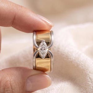 Könnte beinhalten: Ein zweifarbiges Ring mit einem Diamanten im Marquiseschliff. Der Ring hat ein goldenes Band mit silbernen Akzenten und einem funkelnden Diamanten. Das Design ist modern und elegant, geeignet für Verlobung oder Alltag.