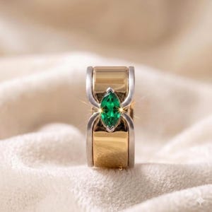 Könnte beinhalten: Ein gold-silberner Ring mit einem smaragdgrünen Edelstein im Marquiseschliff. Der Ring hat ein einzigartiges Design mit einem goldenen Band und silbernen Akzenten. Der Smaragd ist in einer silbernen Lünette gefasst.