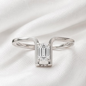 Könnte beinhalten: Silberring mit rechteckigem Smaragd-Diamanten. Der Ring hat ein einzigartiges, geschwungenes Design, das den Diamanten umschließt. Der Markenname "ELANZIA" ist dezent auf dem Band eingraviert. Der Ring ist vor einem weißen, strukturierten Hintergrund platziert.