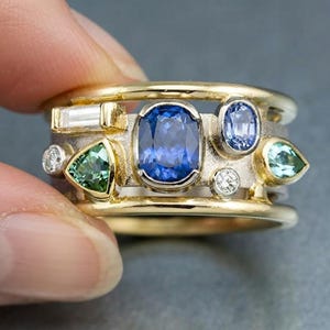 May include: A multi-stone ring featuring a central oval blue sapphire, a baguette-cut diamond, a triangular green gemstone, a pear-shaped light blue stone, and two small round diamonds set on a textured silver band with gold outer rails.