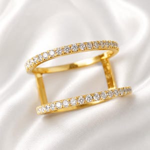 Könnte beinhalten: Ein goldfarbener Doppelbandring mit einer Reihe kleiner, klarer Edelsteine, die entlang der oberen und unteren Bänder gesetzt sind. Der Ring ist vor einem weißen, seidigen Hintergrund platziert, der sein Funkeln und Design hervorhebt.