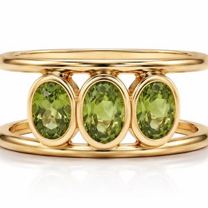 May include: A gold ring featuring a double band design connected by three vertically oriented, oval-cut, light green peridot gemstones. The stones are set in smooth, polished gold bezels, creating a structured and symmetrical appearance.