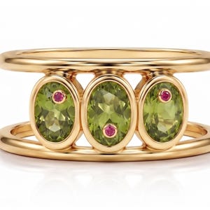 May include: A gold ring featuring two parallel bands connected by three oval-cut, light green peridot gemstones. Each peridot is accented with a small, round, vibrant pink gemstone set directly into the surface of the green stone.