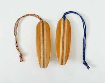 Op de afbeelding: Twee houten bladwijzers in de vorm van een surfplank met een lichtbruin en bruin gestreept ontwerp. Eén heeft een regenboogkleurig koord en de andere een blauw koord. De bladwijzers staan op een witte achtergrond.