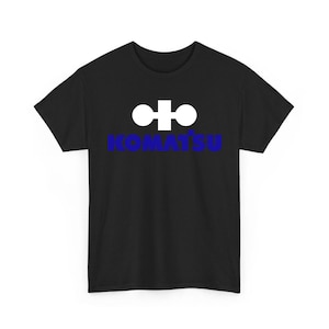 May include: A black short-sleeved t-shirt featuring a white geometric logo above the blue text KOMATSU centered on the chest. The garment has a crew neckline and a standard fit, displaying the brand name and symbol clearly against the dark fabric.