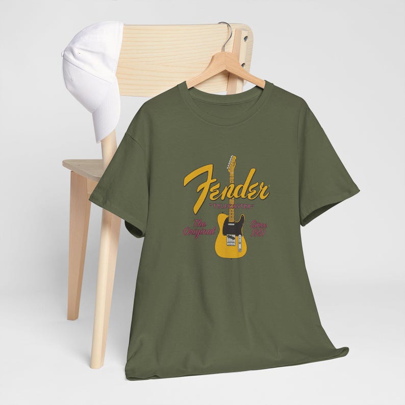 May include: Olive green t-shirt with a Fender guitar graphic. The graphic features a yellow guitar with a black pickguard and the Fender logo in yellow script. The text "The Original" and "Since 1946" are also included.