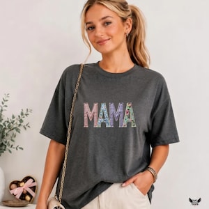 Puede incluir: Camiseta gris oscuro con la palabra "MAMA" en un diseño de patchwork floral colorido. La camiseta tiene mangas cortas y un corte relajado. La imagen muestra a una mujer usando la camiseta.