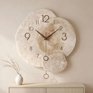 Op de afbeelding: Een decoratieve wandklok met een uniek, gelaagd ontwerp. De wijzerplaat bestaat uit drie onregelmatig gevormde, gebroken witte stukken. Donkerbruine wijzers en cijfers geven de tijd aan. Een kleine, ronde slinger hangt eronder. De diameter is ongeveer 50 cm.