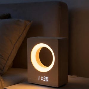 May include: A wooden bedside lamp with a crescent-shaped light and a digital clock displaying 1:30. The lamp is made of light-colored wood and sits on a nightstand. The warm light creates a cozy atmosphere.