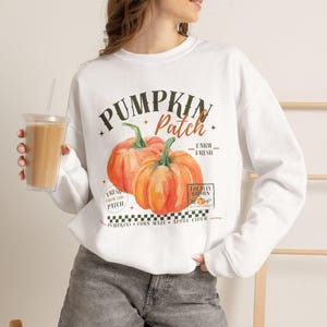 Könnte beinhalten: Ein weißes Langarm-Sweatshirt mit einem Grafikdruck von zwei orangefarbenen Kürbissen und dem Text Pumpkin Patch, Farm Fresh und Locally Grown. Das Design enthält einen schwarz-weißen Karorand sowie die Wörter Pumpkins, Corn Maze, Apple Cider.