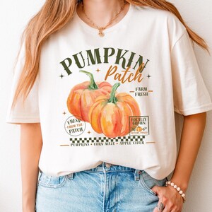 Könnte beinhalten: Ein cremefarbenes Kurzarm-T-Shirt mit einer Grafik von zwei orangefarbenen Kürbissen. Der Text lautet PUMPKIN Patch, FARM FRESH, FRESH FROM THE PATCH, LOCALLY GROWN, AUTUMN PICKED, PUMPKINS, CORN MAZE und APPLE CIDER über einem karierten Rand.