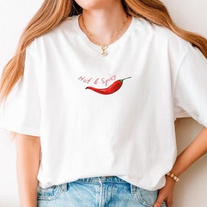 Könnte beinhalten: Weißes T-Shirt mit einer roten Chili-Pfeffer-Grafik und dem handgeschriebenen Text "Hot & Spicy". Das Shirt wird mit hellblauen Jeans kombiniert. Das Gesamtthema ist Freizeitkleidung.