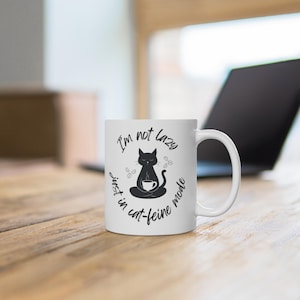 Könnte beinhalten: Weiße Keramik-Kaffeetasse mit einer schwarzen Katzenillustration und dem Text "I'm not lazy just in cat-feine mode". Die Katze ist in meditativer Pose dargestellt und hält eine Kaffeetasse. Die Tasse hat einen gebogenen Griff.