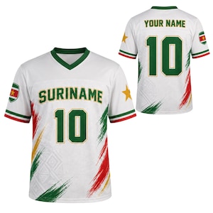 May include: A white V-neck jersey featuring the text SURINAME and the number 10 in green with gold outlines. The design includes green, red, and yellow brushstroke accents, a gold star on the sleeve, and a Suriname flag patch on the shoulder.