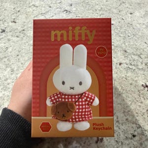 May include: A red box featuring a Miffy plush keychain. The box has the word "miffy" in gold lettering. The plush toy is a white bunny wearing a red and white checkered dress and holding a brown dog. The text "Plush Keychain" is also visible.