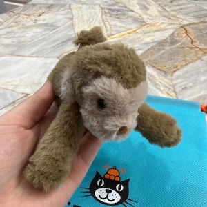 May include: A plush, brown and tan stuffed animal in the shape of a seal. The toy has a soft, furry texture and a small, rounded head. The seal is lying on a blue bag with the text "JELLYCAT LONDON" and a black cat logo.