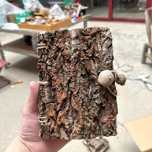 May include: A rectangular journal with a cover made of textured brown tree bark. The spine is bound with light brown twine, and a small wooden acorn-shaped bead is attached to the front cover with a matching twine loop as a closure mechanism.