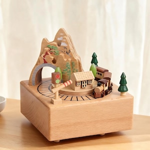 May include: A wooden music box featuring a miniature train scene. The box has a light brown finish and includes a mountain, tunnel, trees, a house, and a train on tracks. The details are intricately carved and painted in natural colors.