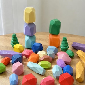 Könnte beinhalten: Eine Sammlung farbenfroher Holz-Bausteine in verschiedenen Formen und Größen. Die Steine sind in Gelb-, Lila-, Blau-, Grün-, Orange-, Rosa- und Rottönen bemalt. Ebenfalls enthalten sind zwei grüne Holzspielzeuge in Baumform.