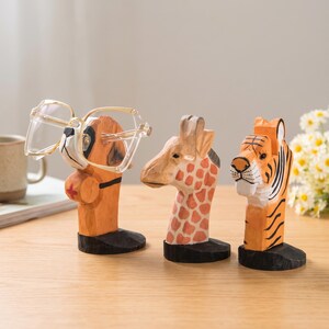 May include: Three hand-carved wooden animal figurines designed as eyeglass holders. The first is a dog wearing a red cross, the second is a giraffe with orange spots, and the third is a tiger with black stripes. Each stands on a black base.