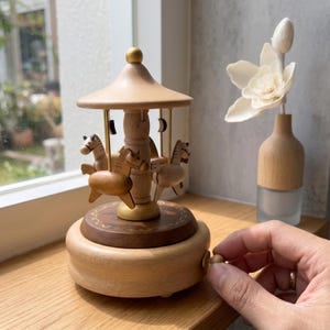 May include: A wooden carousel music box with four horse figurines. The carousel has a light brown, domed roof and a dark brown base with gold accents. The music box is set on a wooden surface near a window, with a decorative flower in a vase.