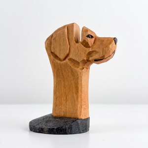 May include: A hand-carved wooden sculpture of a dog head in profile, featuring light brown wood grain with visible chisel marks, a dark brown nose, and a dark, textured base. The sculpture displays a stylized, angular form with a smooth finish.