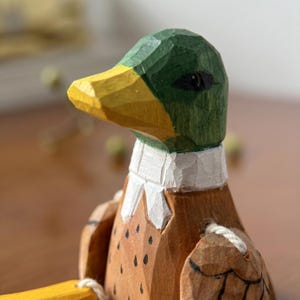 May include: A hand-carved wooden duck figurine featuring a green head, a yellow beak, a white neck ring, and a brown body with black speckles. The figure has visible chisel marks and is attached to a yellow wooden element by a white string.