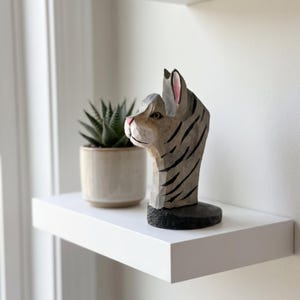 May include: A hand-carved wooden cat figurine featuring a light gray finish with dark gray stripes, a pink nose, and pointed ears. The sculpture is mounted on a circular black base and displays visible wood grain textures from the carving process.