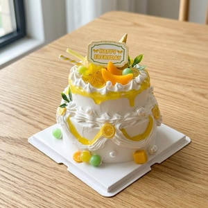 May include: A two-tiered white cake with yellow accents and fruit decorations. The cake is topped with a sign that reads "Happy Everyday". The cake is decorated with star fruit, orange slices, and green candies. The cake sits on a white square cake board.