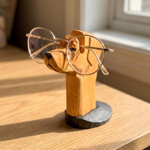 May include: A light brown wooden dog head sculpture serving as an eyeglass holder, featuring carved details and a black circular base. A pair of thin, gold-rimmed round glasses rests securely on the snout of the wooden dog figure.
