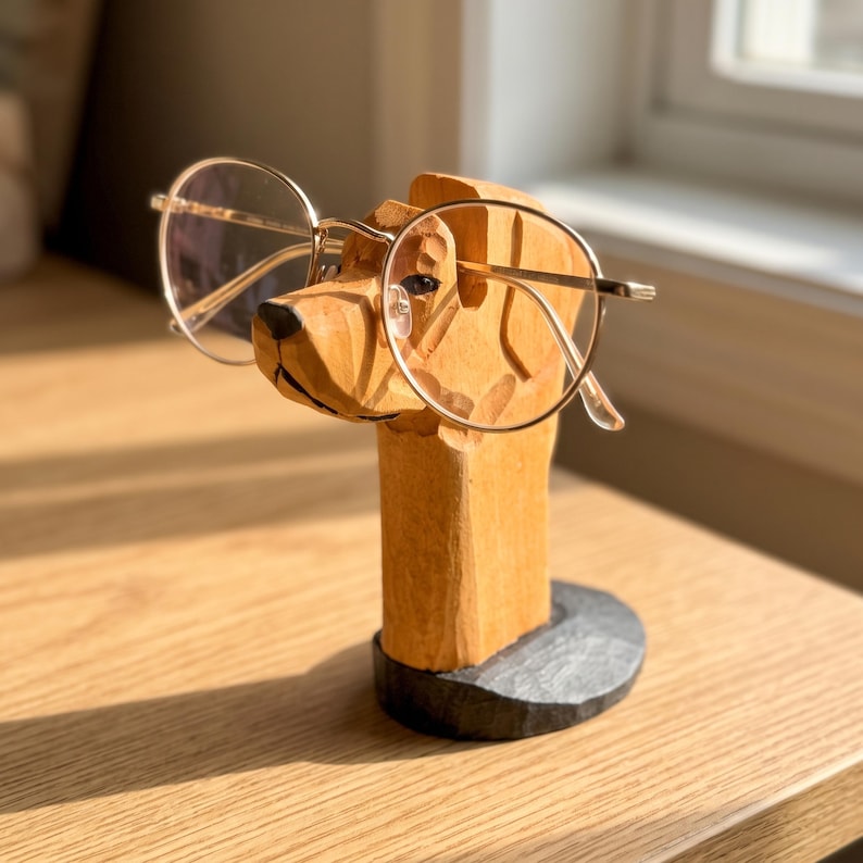 May include: A light brown wooden dog head sculpture serving as an eyeglass holder, featuring carved details and a black circular base. A pair of thin, gold-rimmed round glasses rests securely on the snout of the wooden dog figure.
