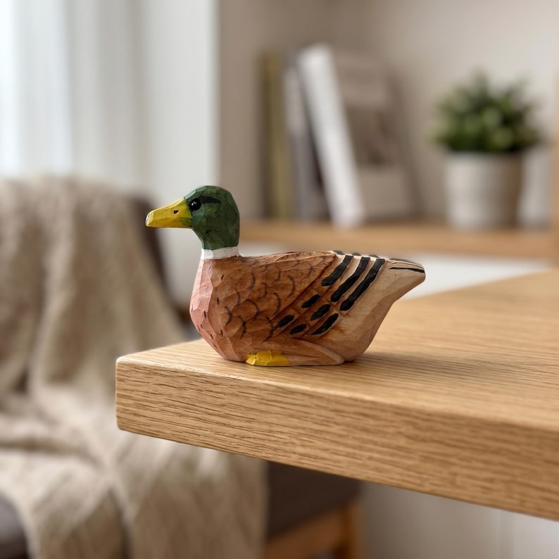 May include: A hand-carved wooden mallard duck figurine featuring a green head, a yellow beak, and a brown body with black wing details. The duck is depicted in a resting position with visible carved textures throughout its surface.