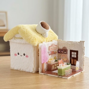 May include: A plush, cream-colored house-shaped toy with a yellow, fluffy roof and a coffee cup accent. The house has a smiling face and the text "FUFU HOUSE". The open side reveals a miniature coffee shop scene.
