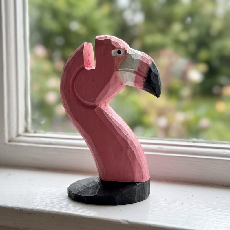 May include: A hand-carved wooden flamingo figurine featuring a pink neck and head with a white and black beak. The sculpture has visible chisel marks throughout its surface and is mounted on a circular black base with a notch at the top.