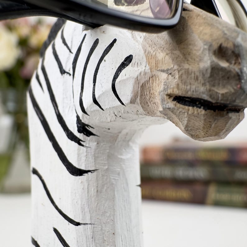 May include: A hand-carved wooden zebra head sculpture painted white with black stripes. The zebra head features a textured, chiseled surface and is wearing a pair of black-framed eyeglasses resting across the bridge of its nose.