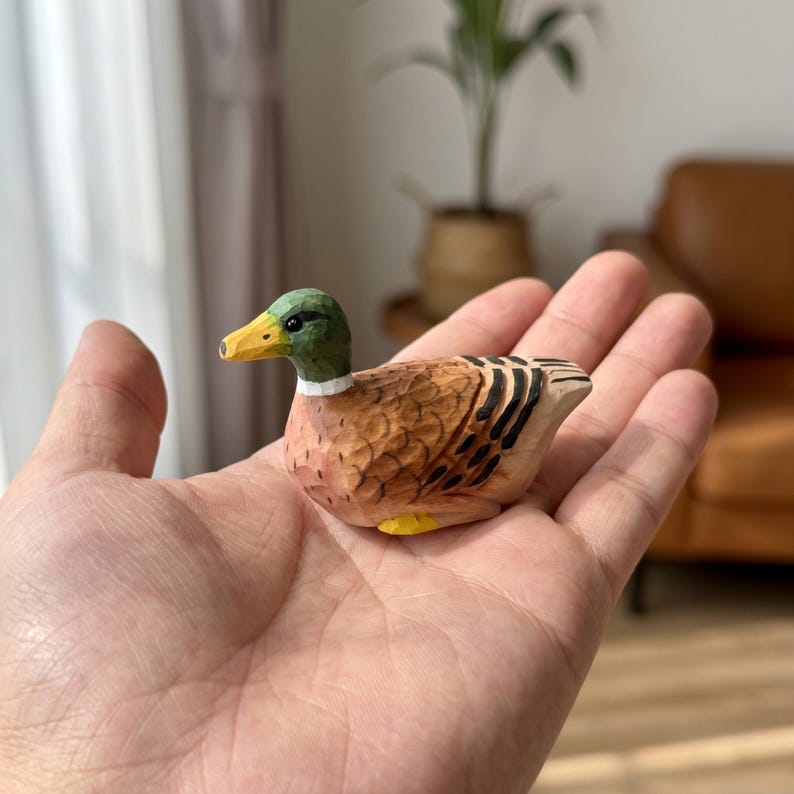 May include: A small hand-carved wooden mallard duck figurine featuring a green head, a yellow beak, and brown textured body feathers with black wing details. The miniature bird is crafted with visible carving marks and painted in natural colors.
