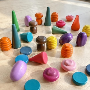 May include: A selection of brightly coloured wooden toys in various shapes, including cones, mushroom-like figures, and rounded shapes. The toys are painted in shades of green, orange, pink, purple, yellow, and blue. The toys are arranged on a light-coloured wooden surface.