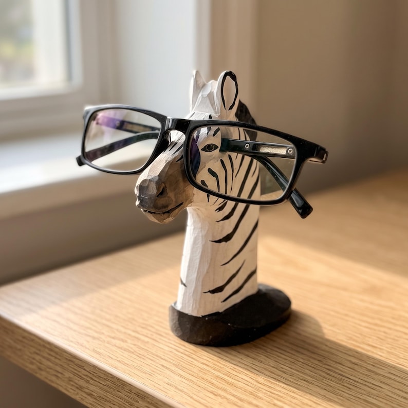 May include: A hand-carved wooden zebra head sculpture designed as an eyeglass holder. The zebra features a white base coat with black painted stripes and a dark oval base. A pair of black-framed rectangular eyeglasses rests on the zebra's snout.