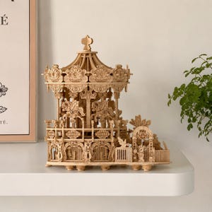 May include: A wooden carousel model with intricate details. The carousel features carved horses, decorative panels, and a domed roof. The model is made of light-colored wood and sits on a white shelf. The carousel is a miniature replica of a classic amusement ride.