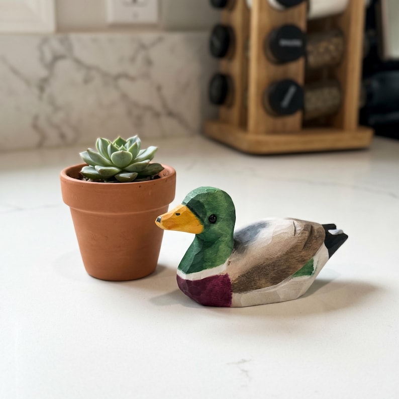 May include: A hand-carved wooden mallard duck figurine featuring a green head, yellow beak, and brown body, positioned next to a small green succulent plant in a terracotta-colored clay pot. The duck has visible carved facets and painted details.