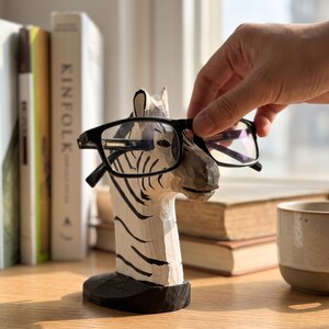 May include: A hand-carved wooden zebra head sculpture designed as an eyeglass holder, featuring a white base with black stripes and a solid black oval pedestal. The zebra head is positioned to support a pair of black-framed eyeglasses on its snout.