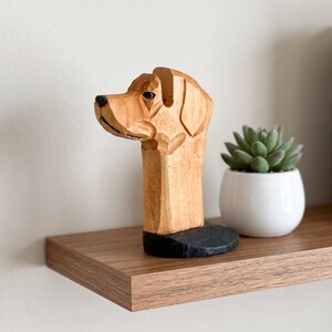 May include: A hand-carved wooden sculpture of a dog head in profile, featuring light brown wood grain, a black painted nose, and a black circular base. The dog has a smooth, stylized finish with visible carved lines defining the ears and snout.